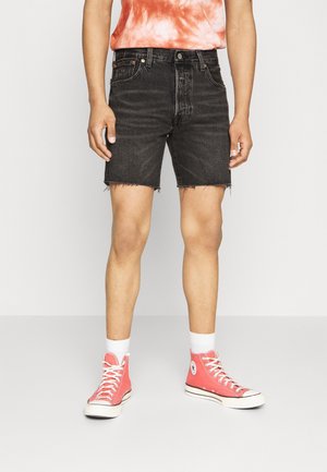 Personne portant un short en denim noir aux ourlets effilochés, des chaussettes blanches, des baskets montantes corail et un t-shirt orange tie-dye rentré dans le short.