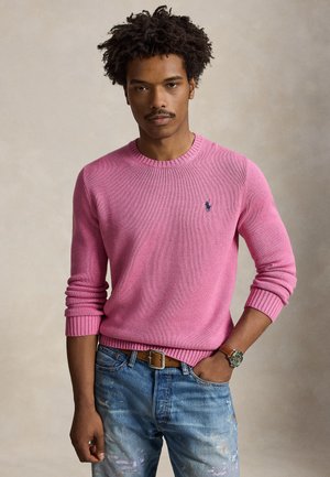Hombre con cabello rizado que lleva un suéter rosa y vaqueros azules, de pie con una mano en el bolsillo frente a un fondo liso.
