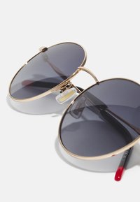 Ochelari de soare rotunzi cu lentile închise, ramă de metal aurie și suporturi de nas transparente. Au un pod dublu subțire și brațe negre cu accente roșii.