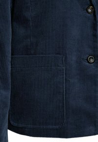 Giacca in velluto a coste blu navy con costine verticali, due bottoni neri e una tasca applicata con superficie testurizzata.