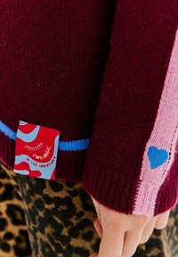 Bordowy dzianinowy sweter z kontrastowymi różowymi i niebieskimi paskami, z niebieskim sercem oraz kolorową metką z napisem "Znajdź magię."