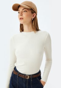 Crème ribbed langsleevetop gecombineerd met hoge taille donkere spijkerbroek, accenten met een bruine riem en een beige baseballcap.