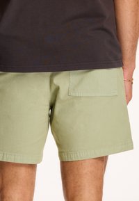 Svetle zelené šortky vyrobené z bavlny, s jednoduchým designem, plochými kapsami a lemovanými okraji. Elastický pas není viditelný.