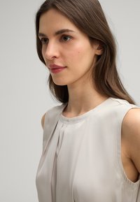 Lichtbeige mouwloze top met geplooid ontwerp, gladde stoftextuur en subtiele stikdetails langs de halslijn en armsgaten.