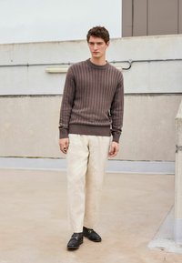 Pull en maille marron, pantalon crème et mocassins noirs. Le pull a un col rond et des poignets ajustés, créant une apparence texturée.