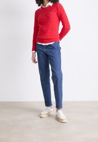 Personne portant un pull rouge à torsades par-dessus une chemise blanche, un jean bleu et des baskets blanches à plateformes, debout sur un sol clair contre un mur blanc.