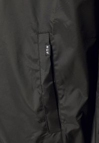 Gros plan d'une poche de veste noire avec une petite étiquette en tissu noire affichant le texte blanc "HUF".