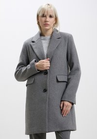 Cappotto grigio in misto lana con un design strutturato, dotato di colletto a lancia, due tasche frontali e bottoni scuri. La texture sembra morbida.