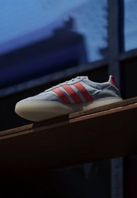 Zapatilla gris con rayas rojas, material superior liso, punta redonda y suela texturizada. Situada sobre una superficie de madera con un fondo oscuro.
