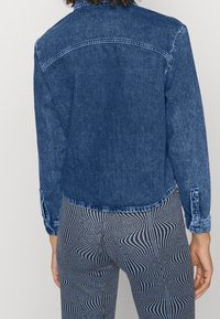 Giacca di jeans blu scuro con orlo curvo, polsini con bottoni e dettagli sulla parte posteriore. Indossata su pantaloni a righe bianche e nere.