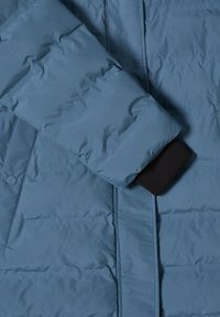 Manga de una chaqueta de invierno acolchada azul con puño ribeteado negro y botón a presión sobre un fondo de tejido texturizado.