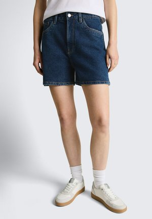 Persoon die donkerblauwe denimshorts van middelmatige lengte draagt, witte sokken, witte sneakers en een wit topje, staand tegen een effen achtergrond.