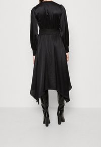 Robe en satin noir avec une taille froncée, des manches longues et un ourlet asymétrique. Portée avec des bottes noires à talons hauts. Texture lisse sur l'ensemble.