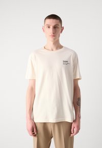Crème-kleurig T-shirt gemaakt van katoen, met een ronde hals, korte mouwen en een zwart logo op de linkerzijde van de borst.