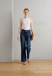 Un débardeur blanc ajusté, un jean en denim bleu foncé taille haute avec des jambes évasées, et des sandales à talons bruns. Tient une pochette brune. Fond uni.