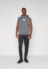 Sivý bezrukávový sveter s bielymi šnúrkami a logom, skombinovaný s čiernymi nohavicami s červeným textom "UFC" a akcentmi, doplnený o čierne tenisky.