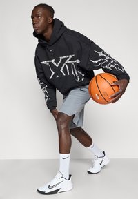 Μαύρο φούτερ με λευκό γραφικό, γκρι σορτς και λευκά αθλητικά παπούτσια με μαύρες λεπτομέρειες. Κρατάει μια πορτοκαλί μπάλα μπάσκετ.