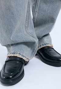 Mocasines de piel negra combinados con jeans de mezclilla azul claro desgastados que presentan dobladillos deshilachados, mostrando un ajuste relajado y una textura suave.