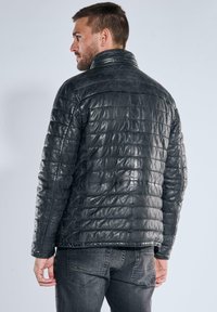 Schwarze gesteppte Steppjacke mit hohem Kragen, einem gesteppte Muster und glänzender Lederoptik. Kombiniert mit dunklen Jeans.