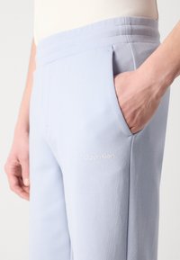 Svijetloplave trenirke s glatkom teksturom, bočnim džepovima i malim bijelim logotipom "Calvin Klein" na butini. Elastična uška.