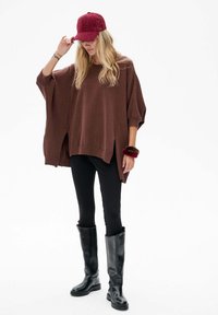 Brązowy oversize sweter z dzianiny z bocznymi rozcięciami, połączony z czarnymi legginsami i wysokimi czarnymi butami. Uzupełniony burgundową czapką i opaską na nadgarstek.