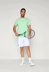 Vaaleanvihreä lyhythihainen urheilupaita, jossa on oransseja yksityiskohtia, yhdistetty valkoisiin shortseihin. Henkilö pitää kädessään tennismailaa.