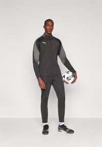 Melns sporta uzvalks ar pelēkiem akcentiem, kas sastāv no jaka ar rāvējslēdzēju un šaurām biksēm. Vīrietis tur melnbaltu rakstu futbola bumbu.