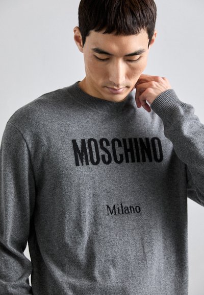 Pilkas megztinis su ryškiu juodu tekstu "MOSCHINO" ir "Milano" žemiau, apvalia iškirpte ir raštuotais rankogaliais, turintis lygią tekstūrą.