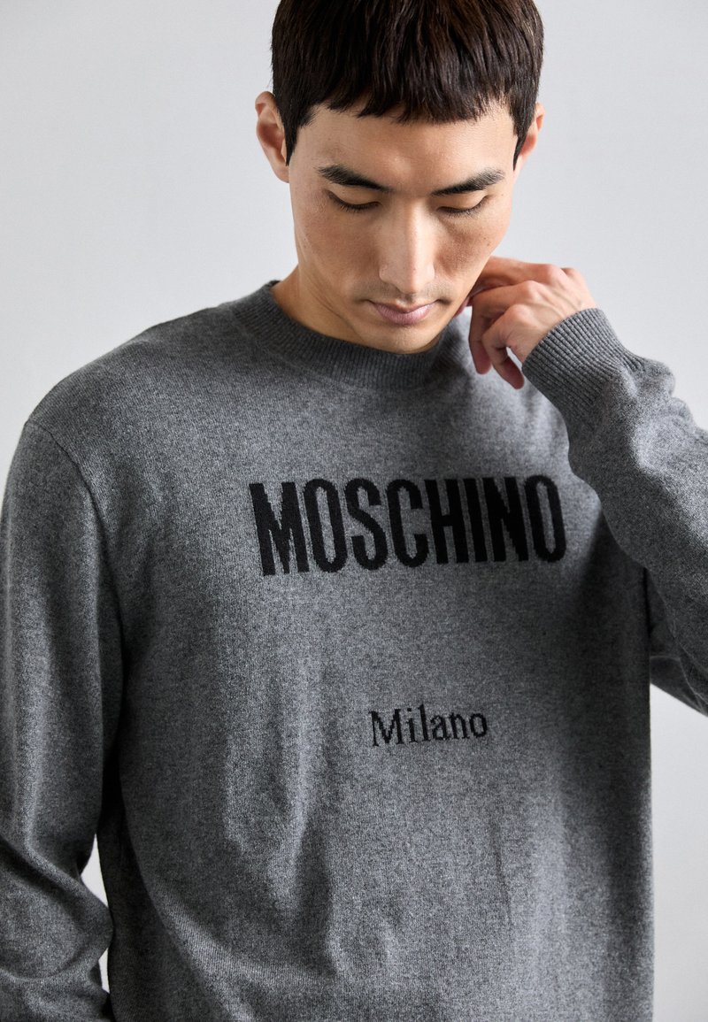 Grauer Strickpullover mit auffälligem schwarzem Text "MOSCHINO" und "Milano" darunter, Rundhalsausschnitt und gerippten Bündchen, mit einer glatten Textur.