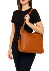 Bruine leren tote bag met een slouchy ontwerp, soepele textuur en een enkele schouderband, vastgehouden door iemand in een zwart shirt en donkere jeans.