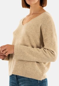 Mujer con un suéter beige de cuello en V, con puños y dobladillo acanalados, combinado con jeans azules, frente a un fondo claro y liso.