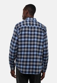 Chemise à carreaux bleue et grise, à manches longues, avec un col boutonné et un ourlet arrondi, présentant des motifs de carreaux contrastants.