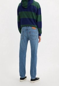 Jeans de denim azul con corte recto, que cuentan con una etiqueta de cuero marrón y bolsillos traseros con detalles de costura. Llevados con una sudadera con capucha a rayas verde y azul marino.