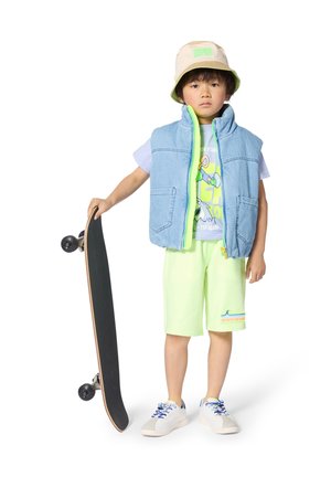 Spijkervest met neon groene ritssluiting, grafisch T-shirt, neon shorts en emmerhoed. Witte sneakers met blauwe veters, met een skateboard in de hand.