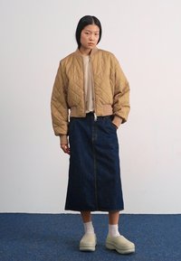 Chaqueta bomber acolchada beige, falda larga de mezclilla oscura y zuecos verdes claros. El fondo es blanco liso con una alfombra azul.