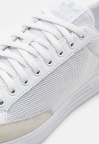 Biezie Adidas kedas ar sietveida augšdaļu, zamšādas akcentiem, plakaniem šņorēm un apaļām caurumiem; minimālisma dizains ar teksturētu detaļu.