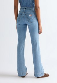 Osoba nosící světlé modré džíny s vysokým pasem a rozšířenými nohavicemi s zadními kapsami a hnědými botami stojí na bílém pozadí.