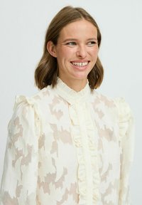 Blusa bianca strutturata con colletto arricciato e chiusura a bottoni. Presenta un motivo floreale e un tessuto trasparente, con maniche corte e una vestibilità rilassata.