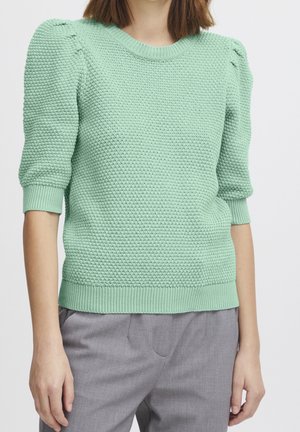 Kvinde iført en tekstureret mintgrøn kortærmet sweater med puffede skuldre og grå skræddersyede bukser, stående foran en ensfarvet baggrund.