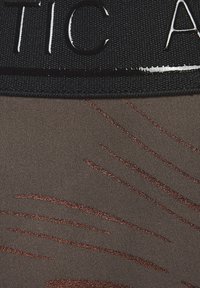 Schwarzes Gummiband mit weißem Schriftzug oben, vor einem glatten braunen Stoff mit metallisch bronzenen wellenförmigen Mustern.