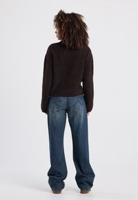 Brauner flauschiger Pullover mit hohem Kragen, kombiniert mit lockeren blauen Jeans aus Denim mit klassischen Gesäßtaschen. Schlichter weißer Hintergrund.