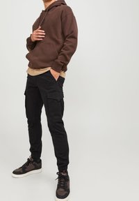 Man med brun huvtröja, beige skjorta under, svarta cargobyxor och svarta och bruna sneakers, står med en hand i fickan.