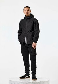 Vyras juoda su gobtuvu striuke, juodomis krovinių kelnėmis ir juodais sportbačiais su baltais detalėmis stovi ant apvalios marmuro platformos.