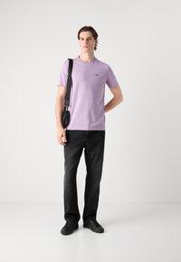 Camiseta de manga corta lila con un logo negro, combinada con jeans negros y zapatillas negras. La persona lleva una pequeña bolsa negra sobre un hombro.
