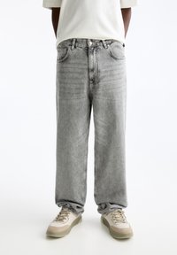 Persona con pantalones vaqueros holgados de color gris claro y zapatillas beige, de pie contra un fondo blanco liso con las manos detrás de la espalda.