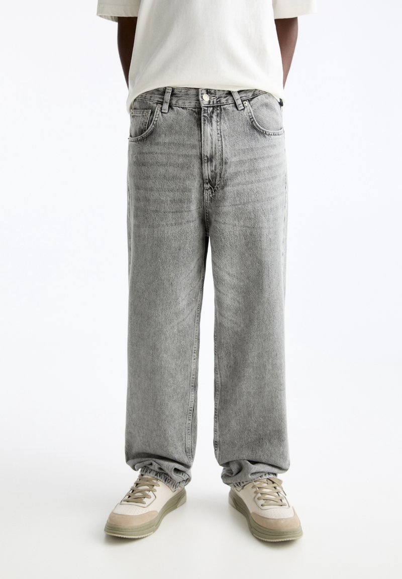 Persona con pantalones vaqueros holgados de color gris claro y zapatillas beige, de pie contra un fondo blanco liso con las manos detrás de la espalda.