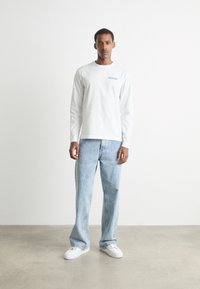 Cămașă albă cu mânecă lungă și un accent dreptunghiular de culoare albastru deschis. Asortată cu blugi denim albaștri deschis și adidași albi, stând pe un podea netedă.