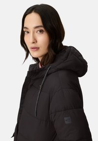 Black puffer jacket with a hood, featuring a zip closure, drawstring accents, and a textured fabric. Logo patch on the left sleeve.
