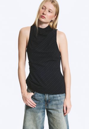 Mujer rubia con un top negro sin mangas y vaqueros azules holgados, de pie con una mano en el bolsillo, mirando ligeramente hacia un lado.
