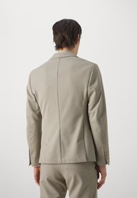 Uomo di spalle che indossa un giaccone sartoriale beige chiaro e pantaloni abbinati, su uno sfondo bianco semplice.